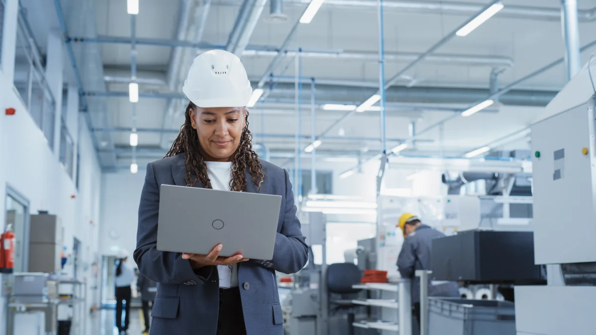 Portrait of a black female engineer in hard hat standing and using laptop computer at electronic manufacturing factory 